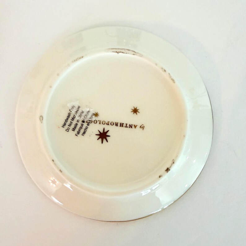 May include: Off-white Anthropologie plate with a gold rim. The base of the plate features a starburst design and the text "by ANTHROPOLOGIE". The plate has a slightly distressed appearance.