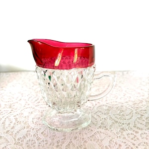 May include: A clear glass creamer with a ruby red rim and handle. The creamer has a diamond-cut pattern on the body and sits on a clear, round base. The creamer is likely vintage or antique.