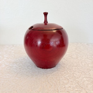 Puede incluir: Un recipiente de madera rojo en forma de manzana con tapa y un pequeño tallo. El recipiente tiene un acabado brillante y se asienta sobre un mantel de encaje blanco. La tapa tiene un pequeño agujero.