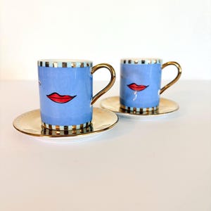 May include: Two blue espresso cups with red lips and gold handles, on matching saucers. The cups have a gold and black striped rim and base. The saucers are gold with a white centre. The cups and saucers are on a white surface.