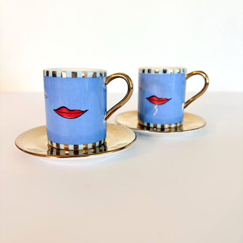 May include: Two blue espresso cups with red lips and gold handles, on matching saucers. The cups have a gold and black striped rim and base. The saucers are gold with a white centre. The cups and saucers are on a white surface.