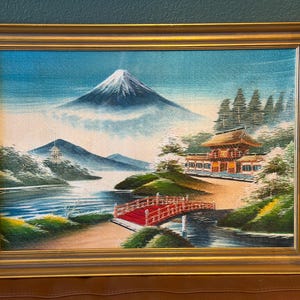 Könnte beinhalten: Gerahmtes Gemälde, das eine japanische Landschaft mit einem schneebedeckten Berg, einer roten Brücke und einem traditionellen Gebäude zeigt. Das Kunstwerk hat einen goldenen Rahmen und zeigt eine ruhige Szene mit blauem Himmel und üppigem Grün.