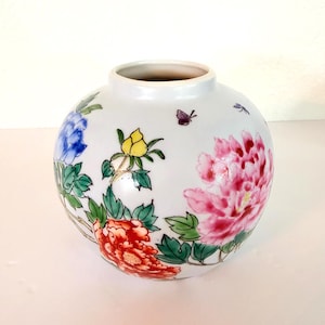 May include: A white ceramic vase with a rounded shape featuring hand-painted floral motifs in blue, yellow, red, and pink. The design includes green leaves, a small purple butterfly, and a tiny blue dragonfly scattered across the surface.