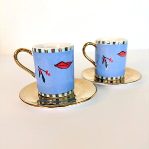 May include: Two blue espresso cups with gold handles and saucers. Each cup features a red lip design and a sprig of berries. The saucers and handles are gold. The cups have a light blue background with a striped pattern.