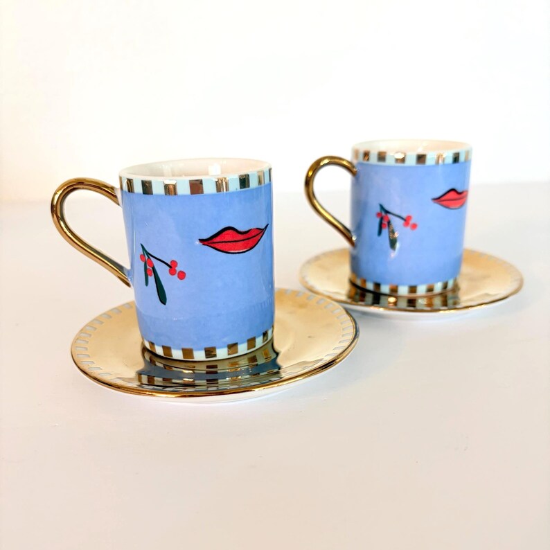 May include: Two blue espresso cups with gold handles and saucers. Each cup features a red lip design and a sprig of berries. The saucers and handles are gold. The cups have a light blue background with a striped pattern.