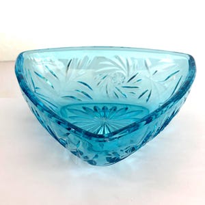 Puede incluir: Un cuenco de vidrio turquesa de forma triangular. El cuenco presenta un patrón floral y de hojas grabado. El borde es liso y redondeado, y el exterior es texturizado. El cuenco está vacío y descansa sobre una superficie blanca.