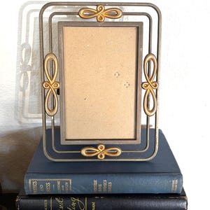 Può includere: Una cornice rettangolare in metallo con un design a fiocco dorato su ogni angolo e al centro. La cornice è appoggiata su una pila di libri con copertine blu e nere. La cornice è vuota.