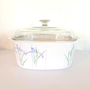Könnte beinhalten: Weißer Auflaufform mit einem Deckel aus klarem Glas. Die Auflaufform hat ein Blumendesign aus lila Iris und grünen Stielen. Die Auflaufform hat einen Knauf aus klarem Glas auf dem Deckel und zwei seitliche Griffe.