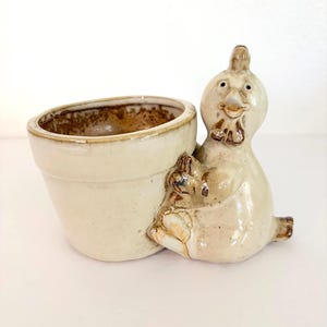 May include: A ceramic planter featuring a cream-colored pot attached to a sculpted chicken figure. The chicken has a glossy finish with brown accents, a small comb, and a beak. The interior of the pot shows a brown, textured glaze finish.