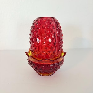 May include: A red glass candle holder with a textured, bumpy surface. The candle holder has a matching base with a yellow rim. The glass is a vibrant red color, and the design is reminiscent of a cactus.
