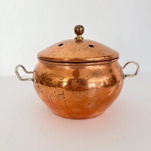 May include: A copper cooking pot with a lid and two handles. The pot has a rounded shape and a shiny, aged finish. The lid has a small knob and two small holes. The handles are made of silver-colored metal.