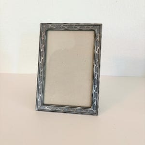 May include: A rectangular picture frame with a metallic silver finish. The frame features a decorative border with a repeating leaf and vine pattern, accented by a beaded edge detail. The center contains a plain, light beige insert.