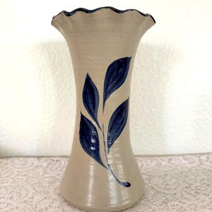 May include: A beige ceramic vase with a flared, ruffled rim and a slender body. The vase features a hand-painted design of blue leaves. The vase is approximately 10 inches tall, perfect for displaying flowers or as a decorative piece.