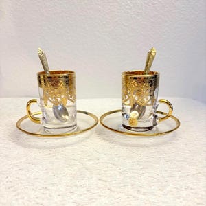 Könnte beinhalten: Zwei klare Teetassen mit goldenen Rändern und Henkeln, jeweils mit passender Untertasse. Die Tassen haben verzierte goldene Filigranmuster und einen kleinen Löffel im Inneren. Das Set steht auf einer weißen Spitzendecke.