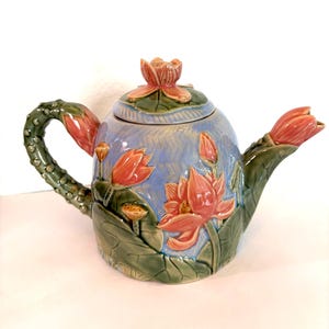 Op de afbeelding: Een keramische theepot met een blauwe body en een bloemenontwerp. De theepot heeft een handvat en tuit versierd met groene bladeren en roze bloemen. Het deksel is bekroond met een roze bloemknop.