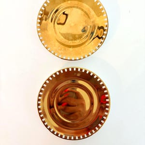 May include: Two gold-coloured decorative plates with a reflective surface. Each plate has a circular shape with a white dotted border. The plates are arranged on a white background, creating a simple and elegant aesthetic.