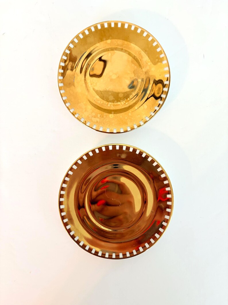 May include: Two gold-coloured decorative plates with a reflective surface. Each plate has a circular shape with a white dotted border. The plates are arranged on a white background, creating a simple and elegant aesthetic.