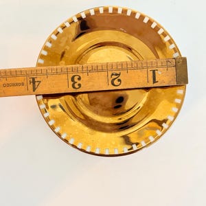 May include: A gold-coloured saucer with a white, rectangular pattern around the rim. The saucer is approximately 10 cm in diameter. A wooden ruler is placed across the saucer for measurement.