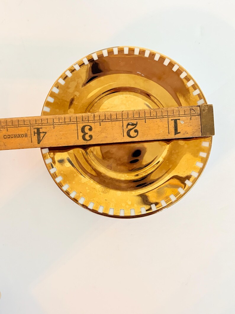 May include: A gold-coloured saucer with a white, rectangular pattern around the rim. The saucer is approximately 10 cm in diameter. A wooden ruler is placed across the saucer for measurement.