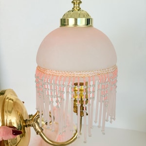 May include: A wall-mounted lamp featuring a polished brass base and a dome-shaped frosted pink glass shade. The shade is adorned with a decorative trim of light pink and clear glass beads that hang in a fringe pattern around the bottom edge.