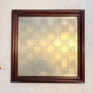 Puede incluir: Un tablero de ajedrez decorativo cuadrado con un marco de madera marrón oscuro. La superficie del tablero es de color dorado claro, dividida en cuadrados con diseños florales grabados. El marco es de un marrón oscuro intenso.