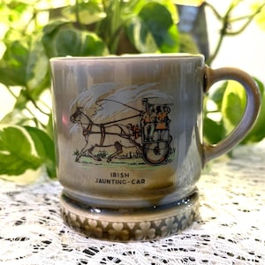 Könnte beinhalten: Graue Keramiktasse mit Henkel und dekorativem Sockel. Die Tasse zeigt eine Illustration eines von Pferden gezogenen Jaunting-Cars mit Passagieren und dem Text "IRISH JAUNTING-CAR". Die Tasse steht vor einem Hintergrund aus grünem Laub.