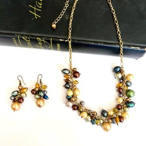 May include: A gold-toned necklace and matching earrings set. The necklace and earrings feature clusters of colorful beads in shades of gold, brown, green, and blue. The jewelry is displayed on a white surface with a dark book in the background.