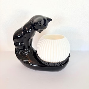May include: A black ceramic cat-shaped candle holder with a glossy finish. The cat curves over a white, ribbed, spherical candle holder. The base is a black, oval-shaped dish. A decorative accent for any room.