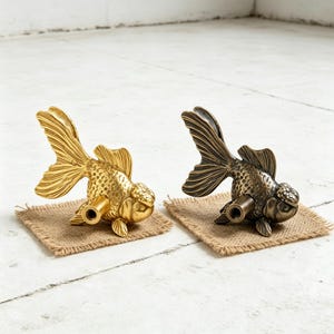 Può includere: Due pomelli decorativi a forma di pesce rosso, uno dorato e l'altro bronzo. Ogni pomello è montato su un piccolo quadrato di juta. I pomelli presentano pinne e squame dettagliate, aggiungendo un tocco di fantasia a qualsiasi mobile.