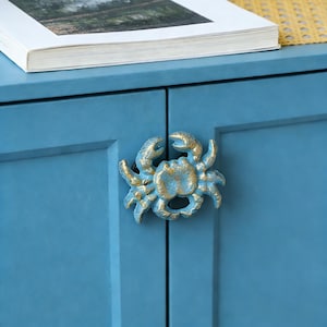 May include: A blue cabinet door with a crab-shaped handle. The handle is painted in a distressed blue and gold finish. A book with a white cover rests on the cabinet.