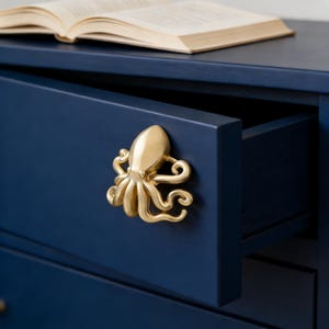 Peut inclure: Tiroir de commode bleu marine avec une poignée dorée en forme de poulpe. Le tiroir est partiellement ouvert, révélant l'intérieur. Un livre ouvert est posé sur le dessus de la commode.
