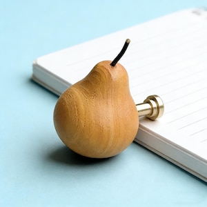 Op de afbeelding: Een houten, peervormige kastknop met een goudkleurige basis en een zwarte steel. De peer is lichtbruin met zichtbare houtnerf. De knop ligt op een wit notitieboek met een lichtblauwe achtergrond.