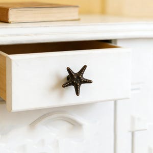 Pode incluir: Gaveta branca com um puxador em forma de estrela do mar em bronze. A gaveta está parcialmente aberta, revelando um interior de madeira. Um livro repousa sobre a superfície acima da gaveta. O puxador adiciona um tema náutico ao móvel.