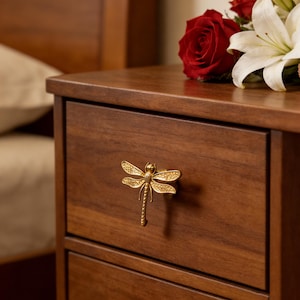 Op de afbeelding: Een close-up van een houten nachtkastje met een decoratieve gouden libelle knop. Het nachtkastje is gemaakt van donkerbruin hout en heeft een gladde, gepolijste afwerking. Rode rozen en witte lelies liggen bovenop.