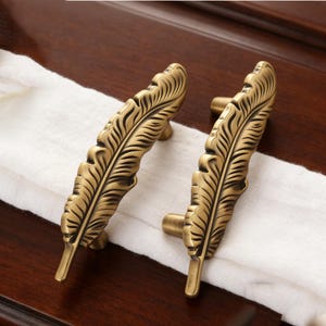 May include: Two antique brass-colored feather-shaped cabinet pulls. The pulls are detailed with intricate feather vein patterns. The hardware is set against a dark wood surface with a white cloth. These decorative pulls add a vintage touch to furniture.