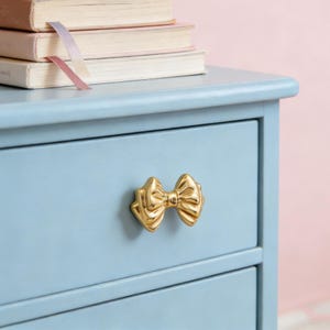 May include: A light blue dresser with a gold bow-shaped drawer pull. The dresser has three drawers and a stack of books on top with a pink ribbon bookmark.