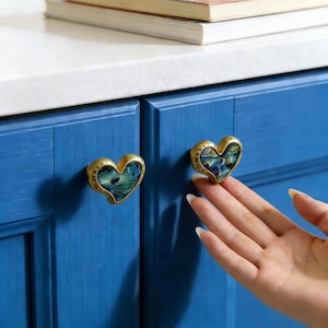 May include: Heart-shaped cabinet knobs with a gold finish and iridescent abalone shell inlay. The knobs are mounted on a blue cabinet door. The knobs are approximately 5 cm wide.
