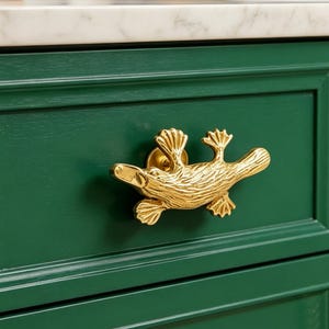 Puede incluir: Un pomo dorado con forma de ornitorrinco está montado en un cajón de armario verde oscuro. El pomo tiene una textura detallada y una pequeña base circular. El cajón del armario está pintado de color verde oscuro y la encimera de arriba es blanca.