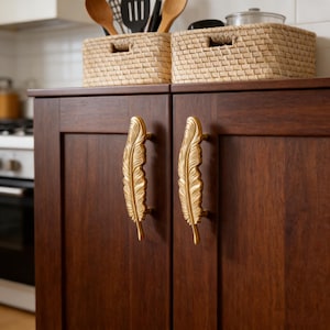 Op de afbeelding: Een donkerbruine kast met twee goudkleurige handgrepen in de vorm van een veer. De kastdeuren zijn gesloten. Twee geweven manden staan bovenop de kast, met keukengerei erin. De keuken heeft een witte achterwand en een fornuis.