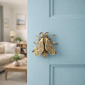 Può includere: Un battente per porta a forma di ape color ottone, montato su una porta azzurra. Il battente presenta ali e segmenti del corpo dettagliati, aggiungendo un tocco decorativo all'ingresso. Lo sfondo mostra un interno sfocato.
