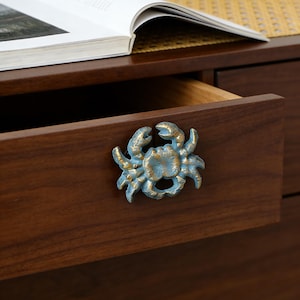 May include: A blue and gold crab-shaped drawer pull is mounted on a dark brown wooden drawer. The drawer is partially open, revealing the interior. An open book rests on a shelf above the drawer.