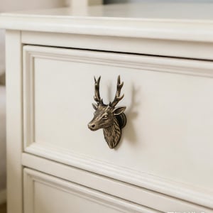 Op de afbeelding: Een bronzen hertenkop-ladeknop is gemonteerd op een wit geschilderde lade. De lade heeft een verzonken paneelontwerp. De hertenkop is gedetailleerd met geweien en gelaatstrekken.