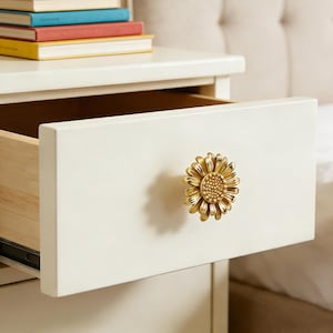 Op de afbeelding: Een witte nachtkast met een open lade met een gouden zonnebloemvormige knop. Bovenop de nachtkast staat een stapel boeken. De binnenkant van de lade is van hout.