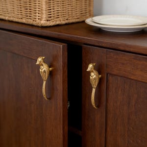 May include: A dark brown wooden cabinet with two doors, each featuring a gold seahorse-shaped handle. The cabinet is topped with a wicker basket and a stack of white plates. The handles provide a decorative accent to the furniture.