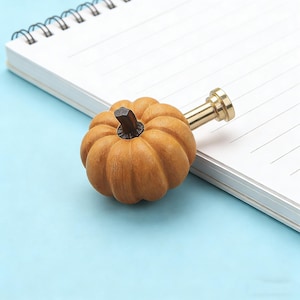 Op de afbeelding: Een oranje pompoenvormige kastknop met een messing steel en een donkerbruine steel. De knop ligt op een wit notitieboek. De knop is een decoratief item met een herfstthema.