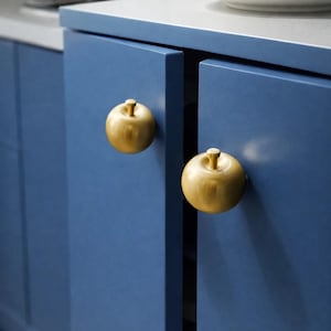 Op de afbeelding: Close-up van blauwe keukenkastdeuren met gouden knoppen in de vorm van appels. De knoppen hebben een warme goudkleur en de kastdeuren zijn effen, matblauw. Witte borden zijn zichtbaar op het aanrecht.