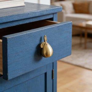 Könnte beinhalten: Ein blauer Holzschrank mit einer geöffneten Schublade. Die Schublade hat einen goldfarbenen, birnenförmigen Griff. Der Schrank hat eine Used-Optik, und ein Stapel Bücher ist oben sichtbar. Der Hintergrund ist verschwommen und zeigt ein Wohnzimmer.