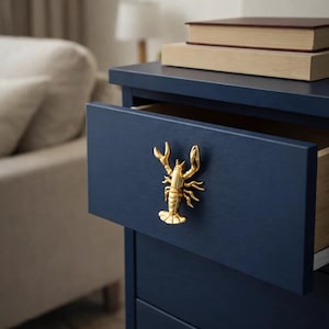 Peut inclure: Une table de chevet bleu marine avec une poignée en forme de homard doré sur un tiroir ouvert. Deux livres sont empilés sur le dessus. La table est dans une chambre.