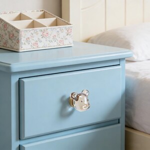 Puede incluir: Una mesita de noche azul claro con un tirador en forma de oso. Una caja de almacenamiento con estampado floral está encima. La mesa está al lado de una cama blanca con una almohada blanca.