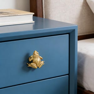 Op de afbeelding: Een blauwe nachtkast met een gouden kikker-vormige ladeknop. De nachtkast heeft twee lades en een boek bovenop. De kikkerknop is gecentreerd op de bovenste lade. De nachtkast staat naast een bed met een wit laken.